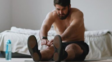 Evde hıyar herifler var. Genç adam vücudunu esnetmeye çalışıyor, yerde oturuyor.