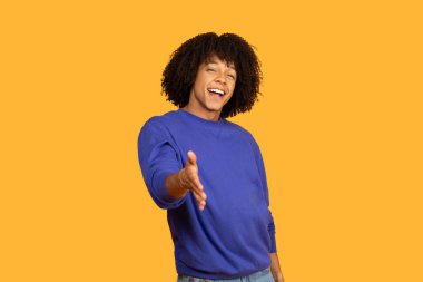 Kıvırcık saçlı, gülümseyen genç bir adam parlak sarı bir arka planda duruyor. Mavi bir kazak ve kot pantolon giyiyor ve elini seyirciye doğru uzatıyor.