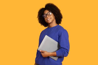 Bu, kıvırcık siyah saçlı, gözlüklü ve mavi kazaklı gülümseyen genç bir adamın fotoğrafı. Önünde gümüş bir dizüstü bilgisayar tutuyor. O, parlak sarı arka planın önünde duruyor..