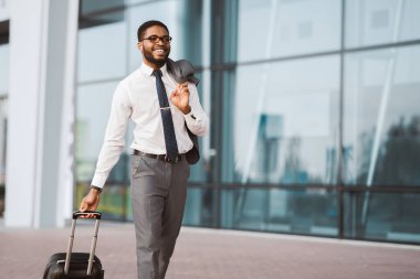 Takım elbiseli Afro-Amerikalı bir adam havaalanında bavuluyla yürüyor. İş seyahatine hazır olduğunu ve profesyonel tavrını vurgulayan, modern ve yoğun bir ortam..