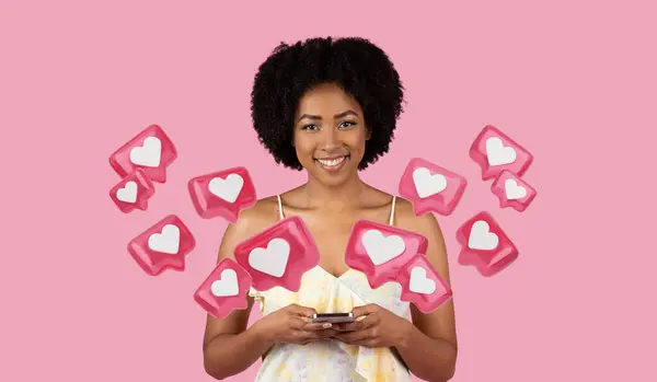 stock image Smiling young African American woman using a cell phone, chatting with guys on dating app, wearing a summer dress against a pink background