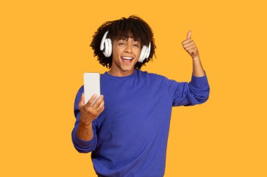 Bu resim, kulaklık takıp telefonuyla müzik dinleyen neşeli bir adamı gösteriyor. Telefonunu sol elinde tutuyor ve sağ eliyle baş parmağını kaldırıyor.