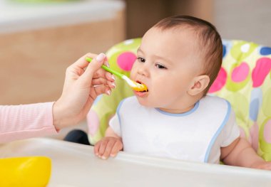 Bir anne, oğlunu mutfak masasında yüksek bir sandalyede otururken besler. Bakımlı ve sevgi dolu bir ortam, sade bir mutfak ortamı..