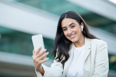 Siyah saçlı, beyaz tişörtlü ve açık renk ceketli genç bir kadın şehir ortamında duruyor. Akıllı telefonunu kaldırırken kameraya gülümsüyor ve kamerayı kullanıyormuş gibi görünüyor..