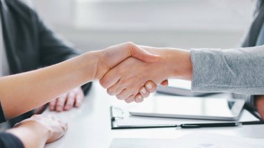 Ofis ortamında toplanan iki iş adamı el sıkışıyor. Belgeler ve arka planda görünür bir kalemle bir masada oturuyorlar.