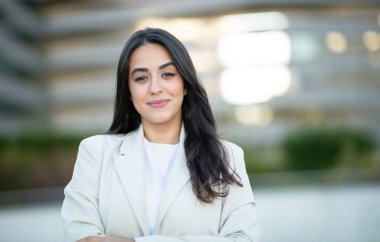Uzun siyah saçlı bir kadın beyaz bir ceket giyiyor ve kendinden emin bir şekilde gülümsüyor. Açık havada duruyor, yeşilliğin bulanık arka planıyla ve bir binayla. Doğrudan kameraya bakıyor..