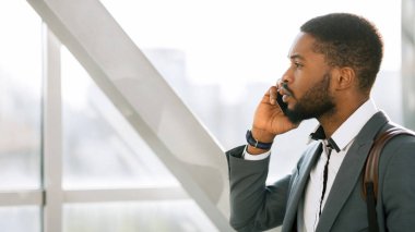 Profesyonel bir siyahi, havaalanının girişinde telefonla konuşuyor. Çevre hareketli ve çağdaş, iş seyahati deneyimini vurguluyor.