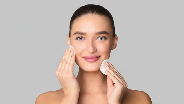 stock image A woman is standing in front of a mirror, applying a facial cleanser to her face with her fingertips. She is gently massaging the cleanser into her skin, focusing on different areas