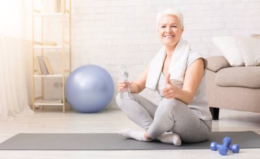 Üzerinde istirahat kadın kıdemli mat sonra fitness egzersiz egzersiz ve başparmak yukarı, şişe su tutarak gösterilen alanı boş