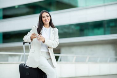 Beyaz ceketli ve pantolonlu genç bir kadın akıllı telefonunu kullanırken gülümseyen bir bavula yaslanıyor. Dışarıda modern bir şehir ortamında duruyor, büyük cam pencereleri ve beton bir cephesi var.