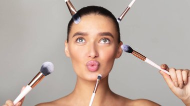 Genç bir kadın, yüzünün etrafına birkaç makyaj fırçasıyla gri bir zemine şakayla poz veriyor. Dudaklarını büzüyor, çeşitli makyaj fırçası uygulamalarını gösteriyor.