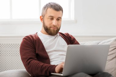 Bir adam kanepede oturuyor, dikkatle dizüstü bilgisayarına odaklanıyor. Çalışırken ya da internette gezinirken elleri klavyede yazmakla meşgul..