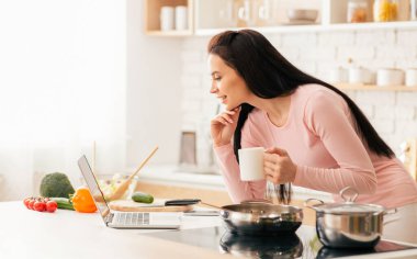 Kadın parlak, modern bir mutfakta elinde kupa tutarken bilgisayarına bakıyor. Taze sebzeler ve mutfak malzemeleriyle çevrili kahvaltı hazırlıyor..
