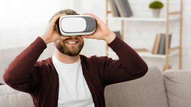 Sanal gerçeklik kulaklığı takan, dijital dünyaya dalmış sakallı bir adam. VR teknolojisiyle etkileşime geçip yeni bir gerçekliği tecrübe ederken odaklanmış ve meşgul görünüyor..