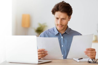 Mavi gömlekli bir adam, elinde laptop ve diğer ofis malzemeleriyle masasında otururken evrakları inceliyor..