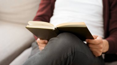 Tanınmayan bir adam kanepeye oturmuş, kitap okumaya dalmış. Kitabı iki eliyle tutuyor, sayfalara odaklanmış, kesilmiş.