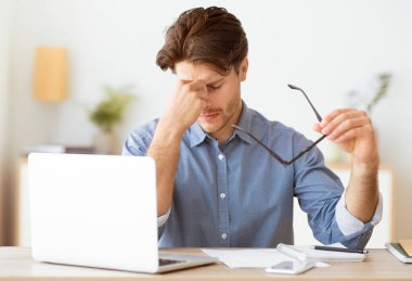 Bir adam masada oturuyor, gözlerini ovuyor ve gözlüklerini tutuyor. Dizüstü bilgisayarı üzerinde çalışırken göz yorgunluğu çekiyormuş gibi görünüyor..