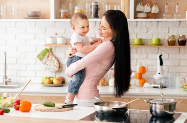 Bir kadın mutfakta durmuş, kucağında bir bebek tutuyor. Kadın yemek pişiriyor ya da hazırlıyor gibi görünüyor ve bebek ona bakıyor. Mutfak aydınlık.