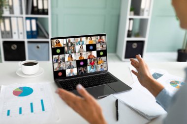 Bir kişi ofis ortamında bir masada oturuyor ve dizüstü bilgisayarında bir video konferansı yönetiyor. Dizüstü bilgisayar ekranında birçok video yayını gösteriliyor, farklı bir grup insan gösteriliyor.