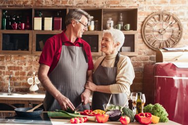 Önlük giymiş yaşlı bir çift mutfaklarında durup birbirlerine bakıyorlar. İkisi de gülümsüyor ve birbirlerine eşlik etmekten hoşlanıyor gibi görünüyorlar. Mutfak rahat ve rahat.