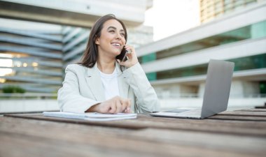 Genç bir iş kadını dışarıda oturuyor, dizüstü bilgisayarı üzerinde çalışıyor ve telefonla konuşuyor. O gülümsüyor ve işinden hoşlanıyor gibi görünüyor. Arkaplanda geniş pencereli bir ofis binası yer alıyor.