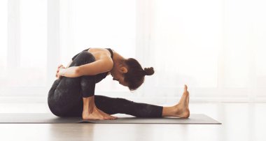 Parlak bir stüdyoda bir kadın yoga pozu veriyor. Bacaklarını ve sırtını esnetiyor, esnekliğini ve odaklanmasını gösteriyor..