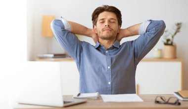 Bir adam dizüstü bilgisayarı ile masasında oturuyor, gözleri kapalı ve elleri başının arkasında, dinlenmek için zaman ayırıyor..