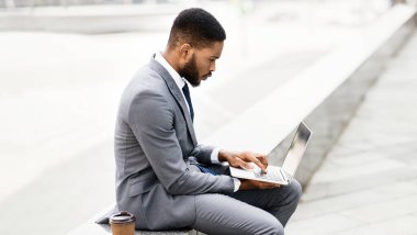 Afro-Amerikalı iş adamı, dışarıda, şehir ortamında otururken bilgisayarını kullanarak kahve içiyor.