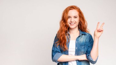Canlı kızıl saçlı bir kadın, barış işareti jesti yapmak için elini kaldırıyor, uyum ve birliği simgeliyor. Günlük kıyafetler giyiyor ve kendinden emin ve etkileyici görünüyor..