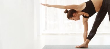 Bir kadın kapalı bir yerde yoga pozu veriyor. Kolları açık bir şekilde tek bacağa odaklanmış ve dengede duruyor. Oda aydınlık, ve o sakin ve minimalist bir ortam ile çevrili.