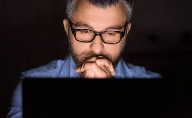 Gözlüklü bir adam, loş bir odada dizüstü bilgisayarı üzerinde çalışırken dikkatle konsantre oluyor. Ekrana odaklanırken elleri çenesinin altında birbirine yapışmış..