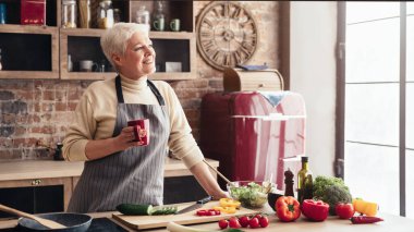 Son sınıf öğrencisi bir kadın mutfağında taze salata hazırlıyor. Bir elinde bir kupa tutuyor ve diğer eliyle bir kase salata karıştırıyor. Mutfak modern ve geniş pencereli.