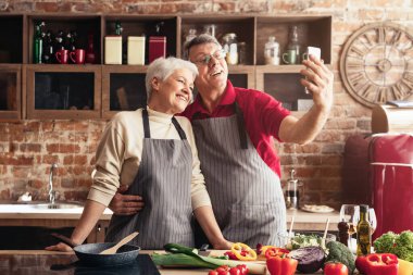 Yaşlı ve mutlu bir çift restoran mutfağında selfie çekiyor. Kadın onun yanında dururken, adam telefonu tutuyordu. İkisi de gülümsüyor ve eğleniyor gibi görünüyorlar..