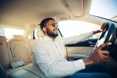 Sakallı ve güneş gözlüklü Afro-Amerikalı bir adam beyaz gömlek giyerken araba kullanıyor. Arabayla gezmekten hoşlanıyor gibi görünüyor, pencereden dışarı bakıyor ve gülümsüyor..