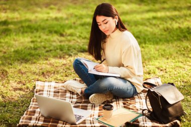 Asian young woman sits on a blanket in a sunny park during the afternoon. She is focused on writing in a notebook, with a laptop, coffee cup, and backpack nearby, studying or working on a project. clipart