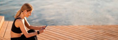 Açık havada antrenman yaptıktan sonra akıllı telefon kullanan orta yaşlı sporcu kadın, Nehir kenarındaki ahşap iskelede dinlenirken cep telefonunda gülümseyen atletik bayan mesajlaşıyor, Boşluğu Kopyala