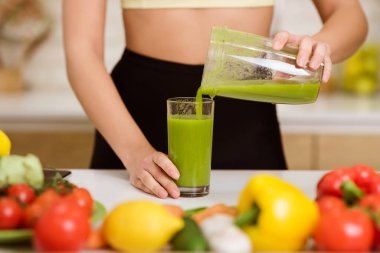 Bir kadın mutfak tezgahındaki taze ürünlerle çevrili bir bardağa yeşil smoothie dolduruyor..