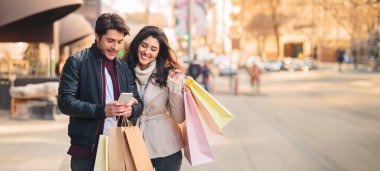 Genç çift yeni butik arıyor, akıllı telefon açık havada kullanarak, kağıt torbalar ile yürüyüş, boş alan