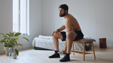 Ev antrenmanı. Çıplak gövdeli genç adam dambılı kaldırıyor, sandalyeye oturuyor.
