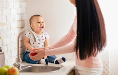 Neşeli bir bebek mutfak tezgahında oturur, annesi lavaboda kırmızı bir elma yıkarken kıkırdar, sabah ışığında yıkanır. Sahne, sevgi ve mutlulukla dolu iç açıcı bir etkileşim yakalar..
