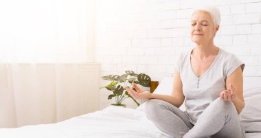 Sakin ve nefes alın. Yatakta Lotus pozisyonda meditasyon kıdemli kadın, Padmasana, boş alan