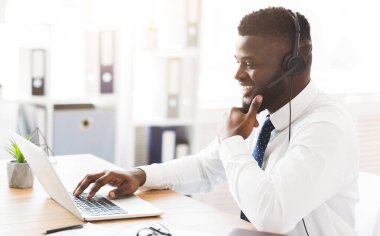 Kulaklıklı neşeli, genç Afrikalı işçi ofiste dizüstü bilgisayarın önünde oturuyor.