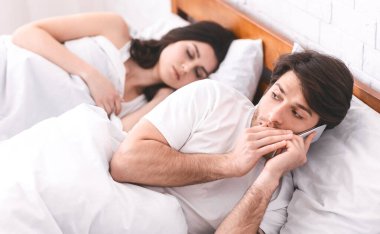 Bir adam yatakta yatıyor, kız arkadaşı yanında uyuyor, o da gizlice telefonda konuşuyor. Adamın eli ağzını kapatıyor ve ifadesi sessiz olmaya çalıştığını gösteriyor..