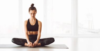 Huzurlu genç bir kadın lotus pozisyonunda yoga minderinin üzerinde gözleri kapalı oturuyor. Geniş pencereli iyi ışıklandırılmış bir stüdyoda ve yumuşak sabah ışığı sakin atmosferi vurguluyor.