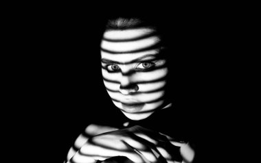 Bu siyah ve beyaz görüntü çizgili bir nesnenin gölgeleriyle kaplı bir kadının yüzünü gösteriyor. Nesnenin ışığı yüzünde yatay çizgiler oluşturarak yüzün özelliklerini gizliyor..