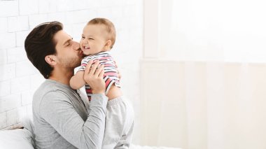 Bir baba, küçük kızını kucağında tutuyor ve ona gülümserken yanağından öpüyor. Baba beyaz çarşaflı bir yatakta oturuyor. Arkasında beyaz tuğla bir duvar var..