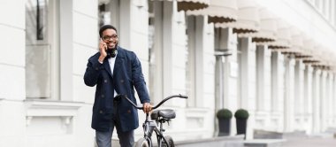 Afrika kökenli Amerikalı bir işadamı dışarıda bisikletin üzerinde otururken telefonla konuşuyor. Şehir ortamında geçen dinamik ve profesyonelce bir ortam..