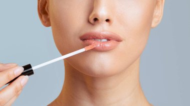 Düz derili bir kadın, dudaklarına parlak bir aplikatör değneği sürüyor. Arka plan açık mavi, kırpılmış.