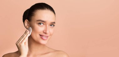 Bir kadın aynanın önünde duruyor ve parmak uçlarını kullanarak yüzüne yüz temizleyici sürüyor. Temizleyiciyi nazikçe cildine masaj yapıyor. Dairesel hareketlerle uzayı temizliyor.