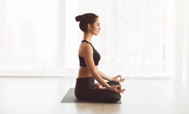 Bir kadın yoga pozisyonunda oturuyor, gözleri meditasyonda kapalı. Yoga yaparken odaklanmış ve huzurlu görünüyor..